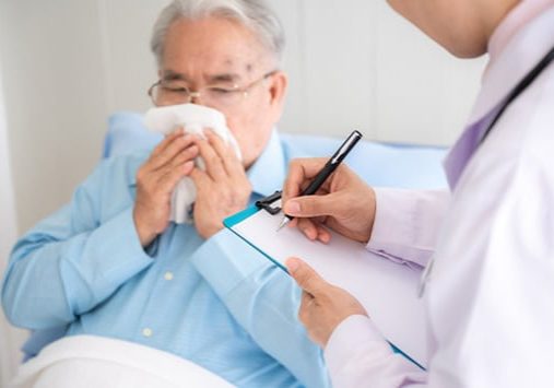 Doctor taking notes while elderly man blows his nose in bed.