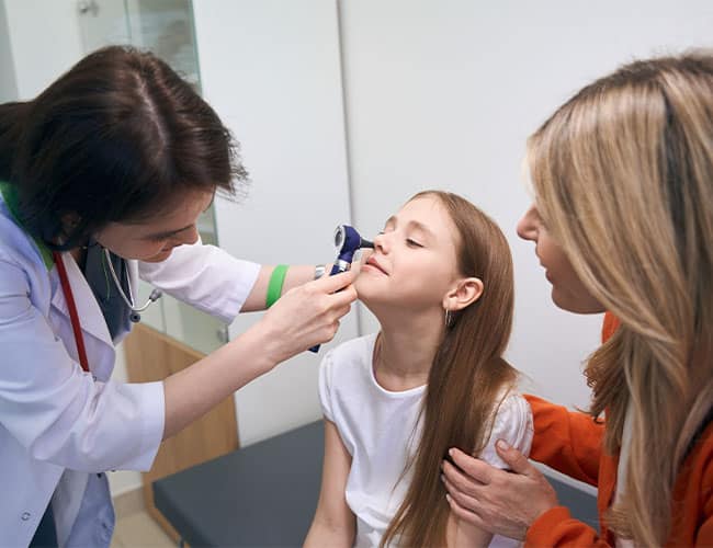 Doctor examining a young girl's nose with a medical device as her mother watches.